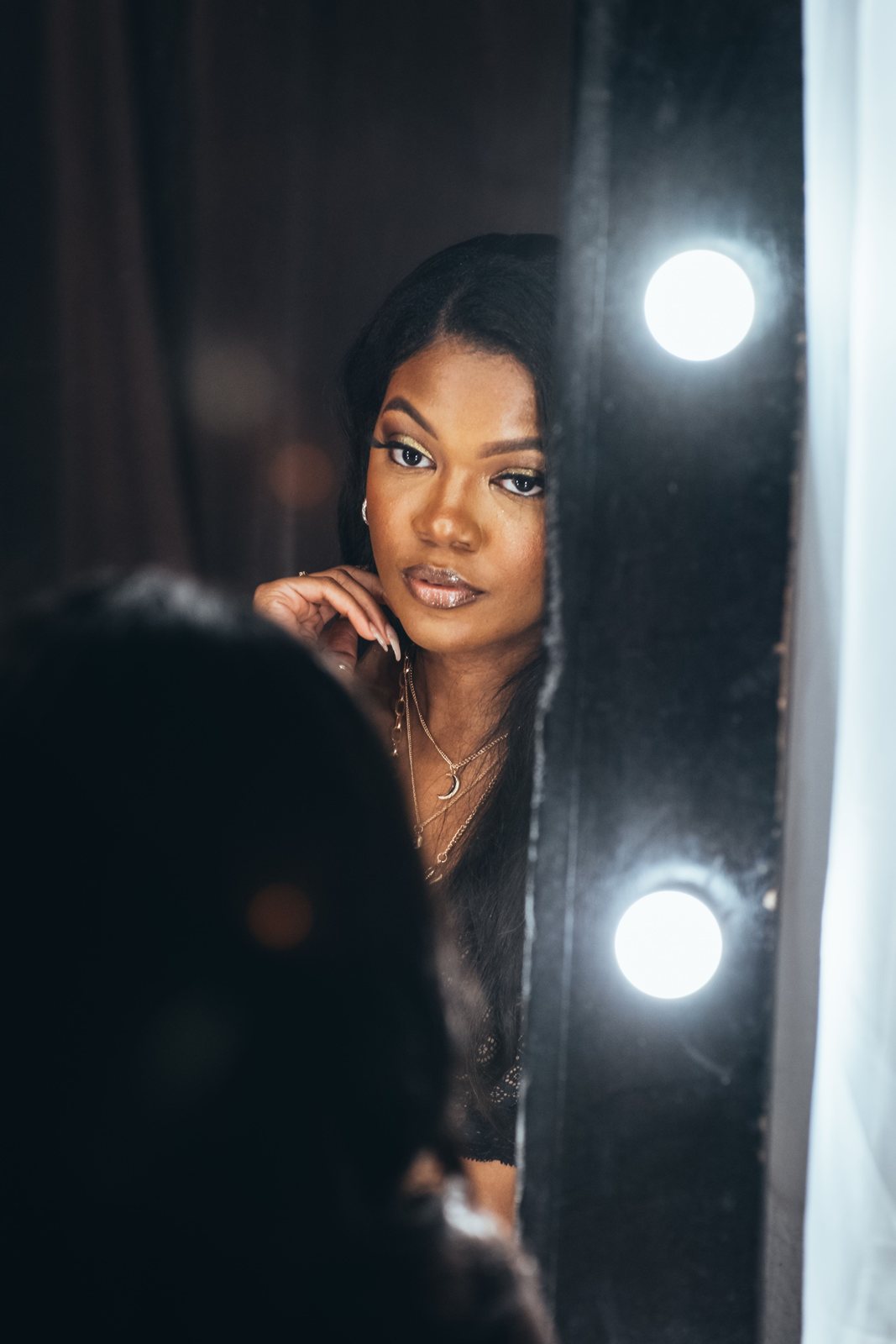 A woman looking out from behind a dark curtain, two round lights beside her.