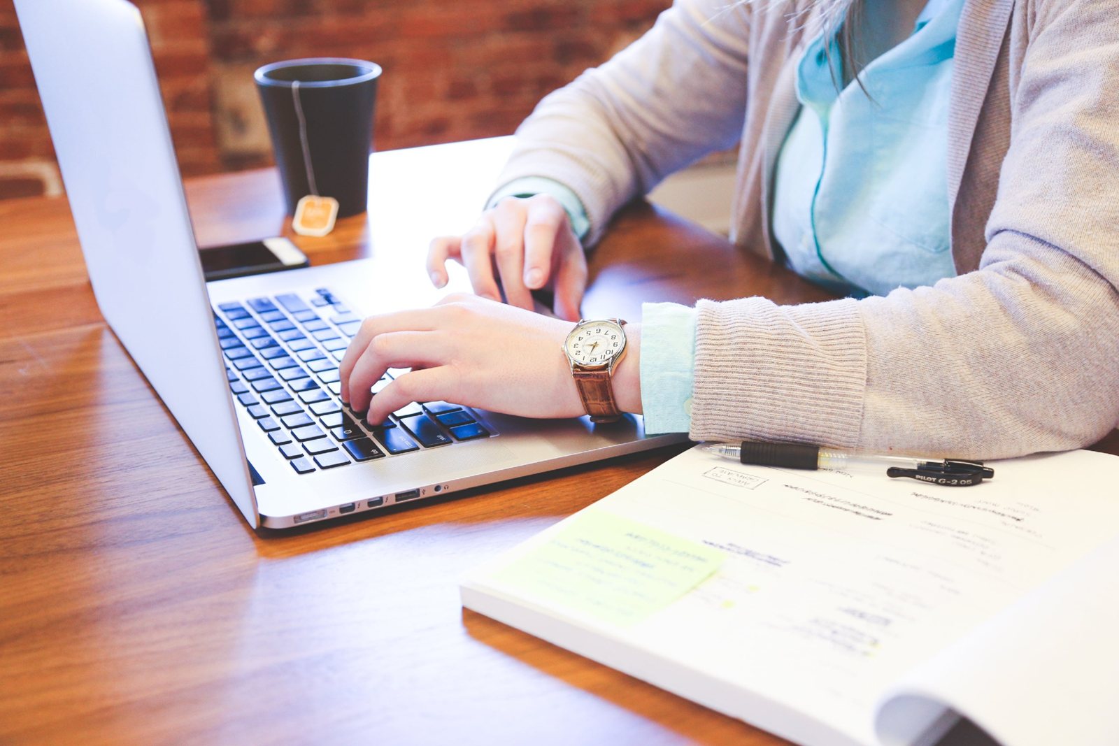 Someone typing at a laptop at a wooden table, an open notebook and a coffee cup beside them.