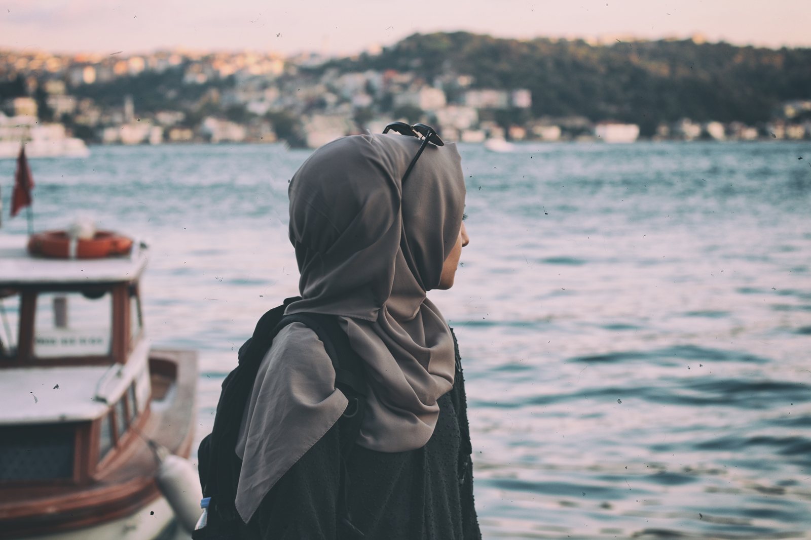 A woman in a grey headscarf looking out across water towards a hillside city, a moored boat behind her.