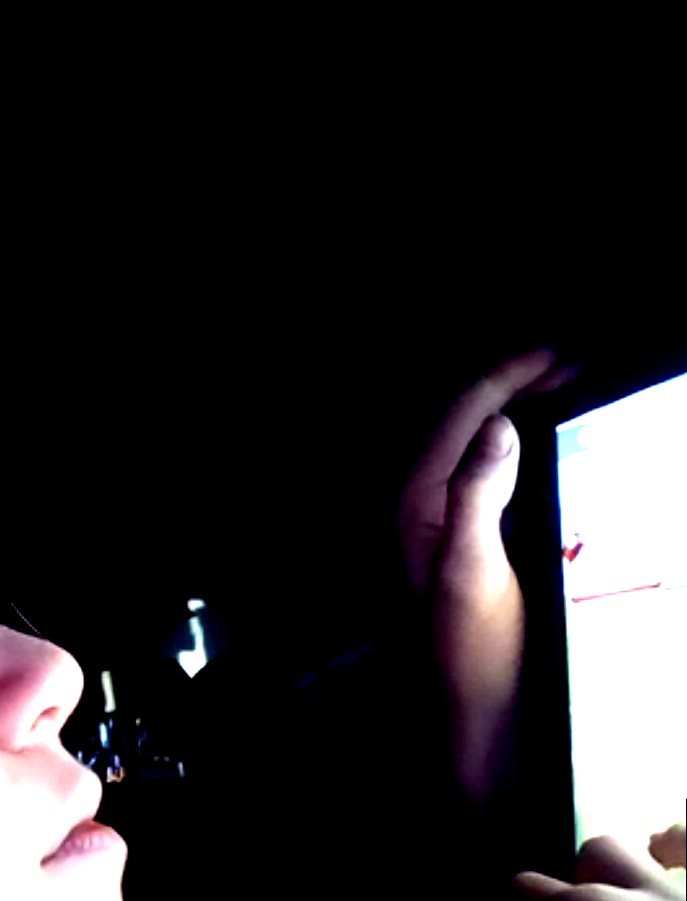 In near-darkness, a small flame at a person’s lips and a hand holding a lit screen.