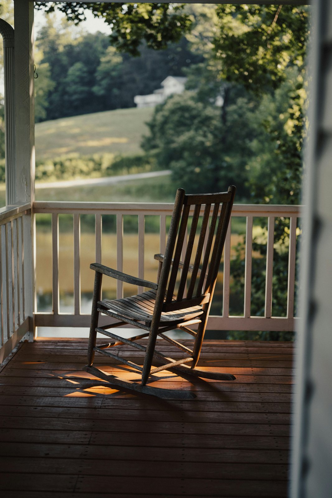 A wooden rocking chair on a porch looking out to a green field and trees.