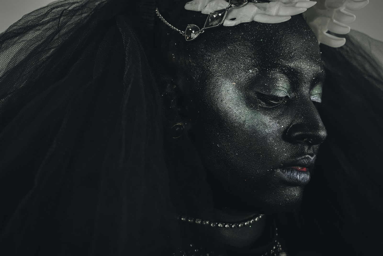 A dark-toned portrait: a person in a black feathered wrap, head turned away, pale hands at their hair.