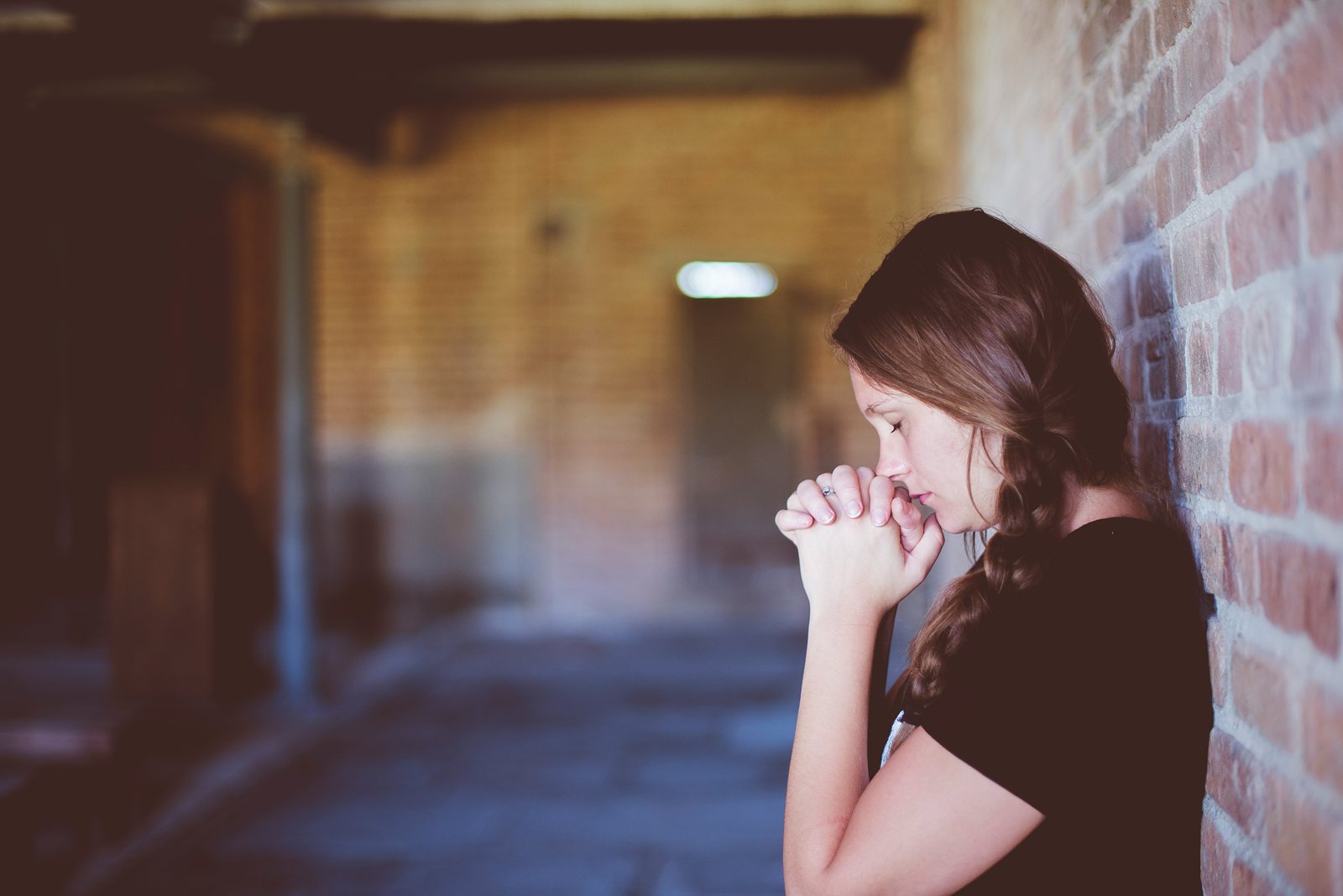 A girl leaning against a brick wall in a corridor, hands clasped at her mouth.
