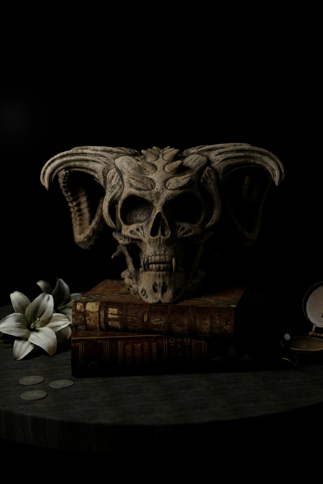 A three-faced skull ornament on a stack of old books beside a white lily.