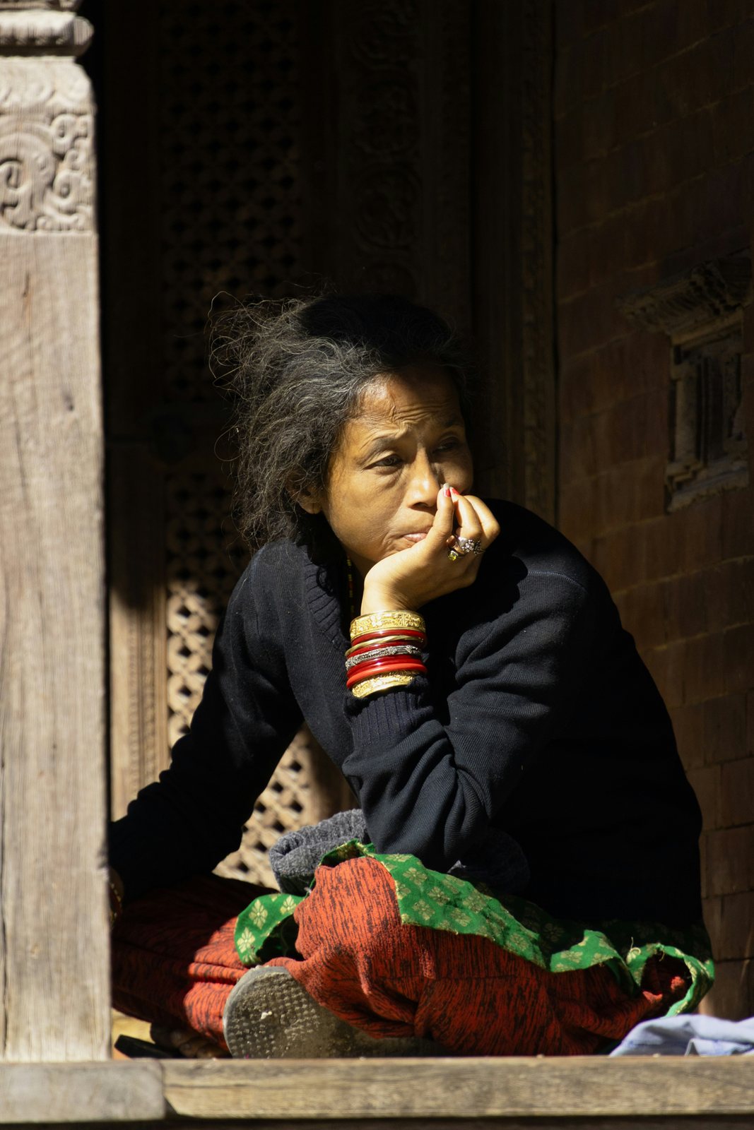 An older woman resting her chin on her hand in a carved stone doorway, bundles of cloth beside her.