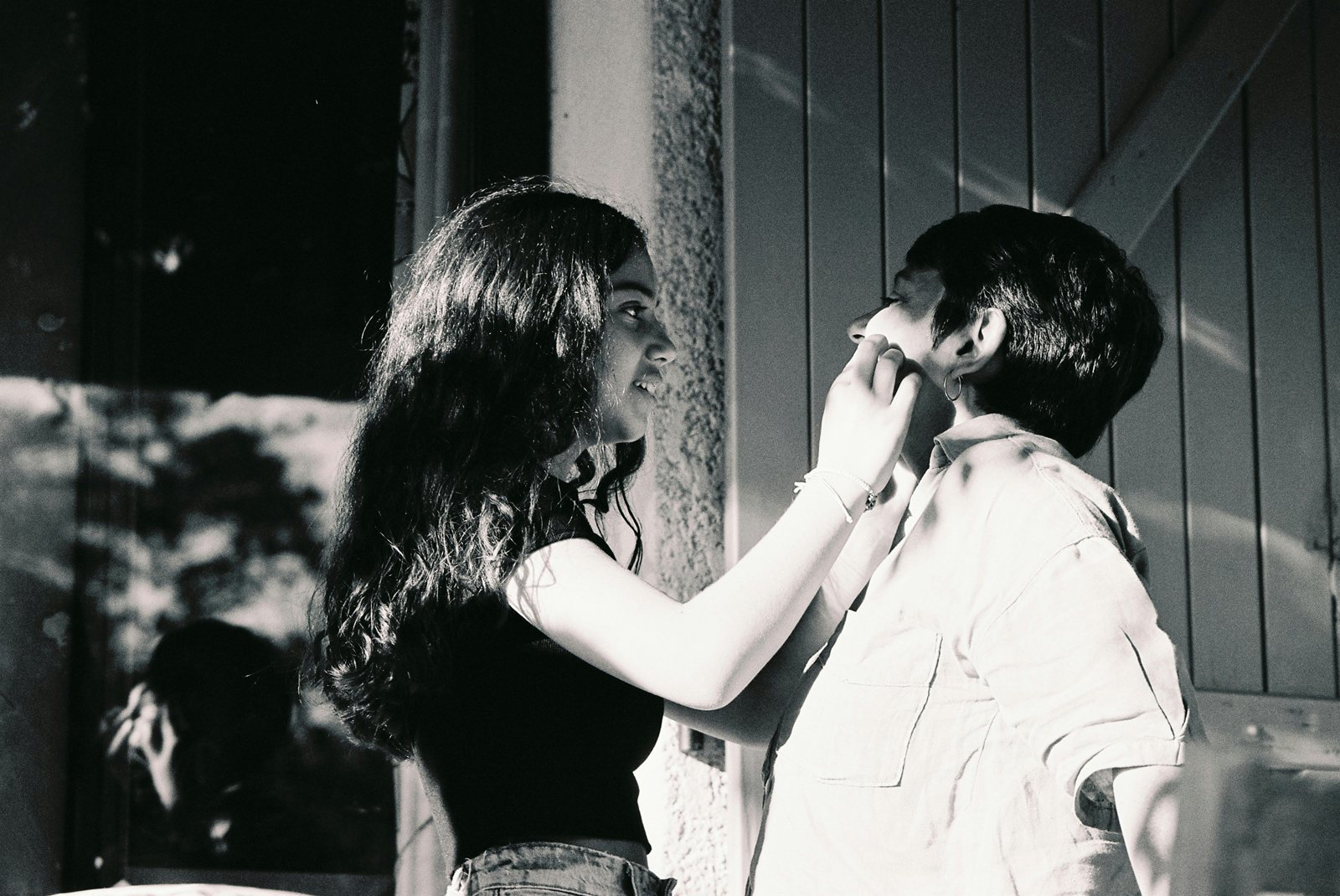 A black-and-white photograph of two young women face to face on a porch, one touching the other’s cheek.