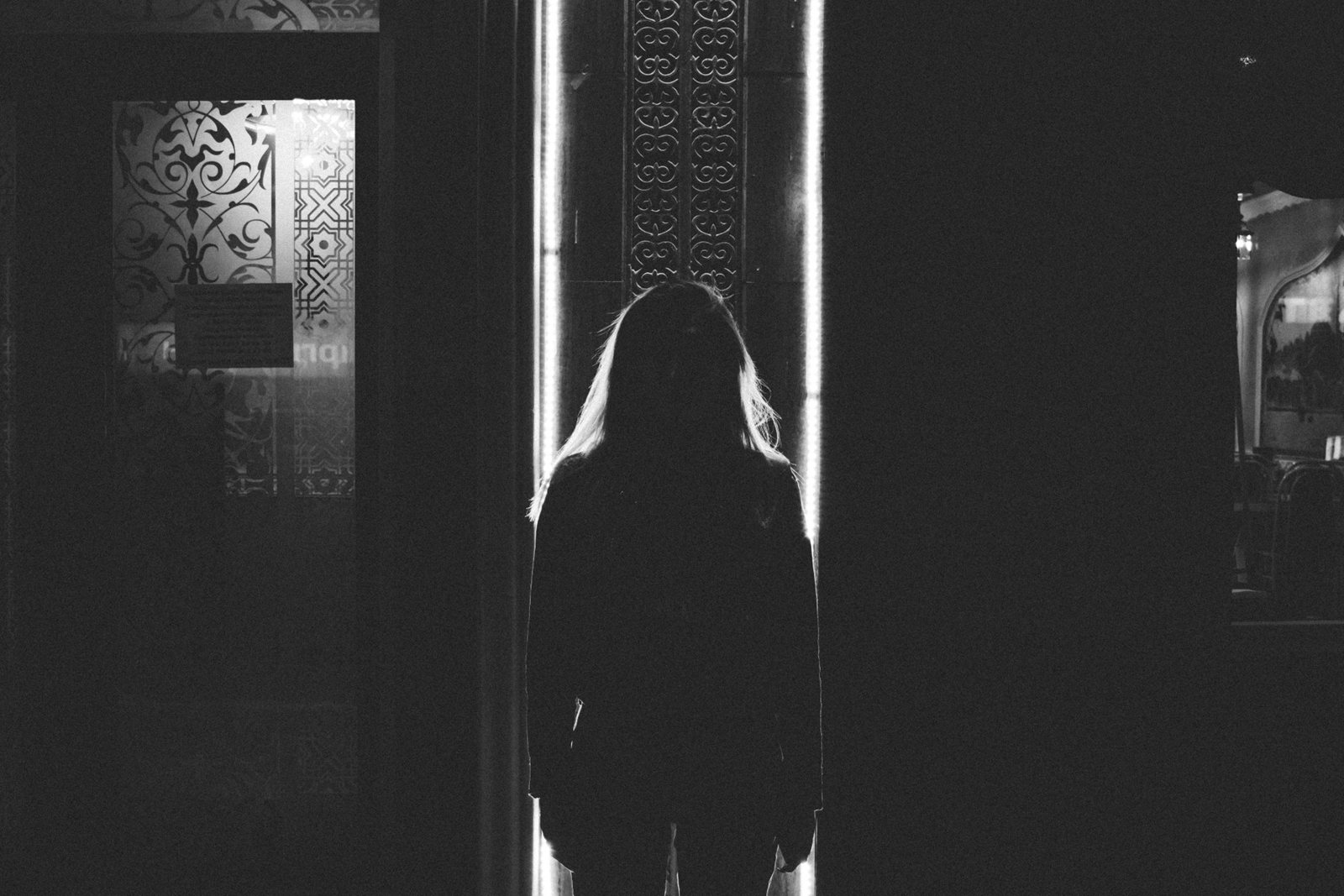 A long-haired figure standing in a narrow lit doorway at the end of a dark room, in black and white.