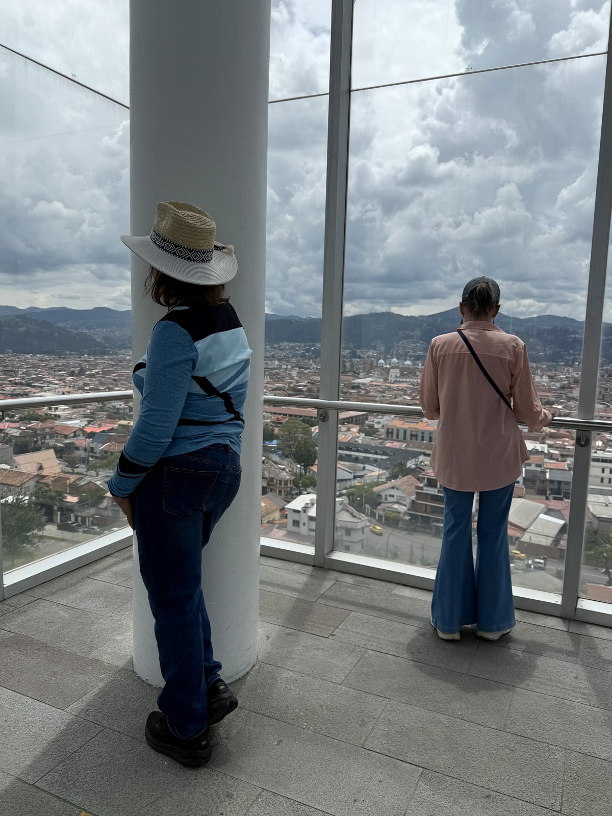 Two people at a high window looking out over a city and distant hills under heavy cloud.