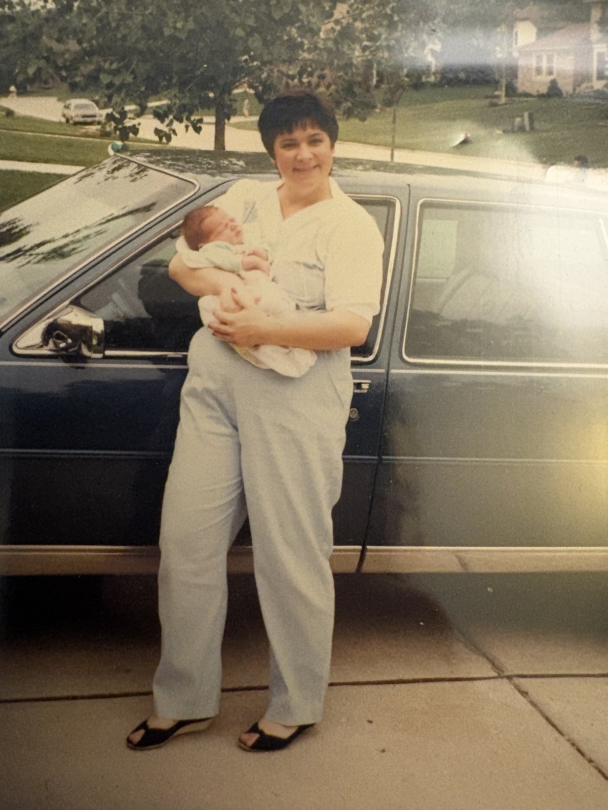 An old photograph of a woman in white trousers holding a newborn beside a parked car on a suburban driveway.