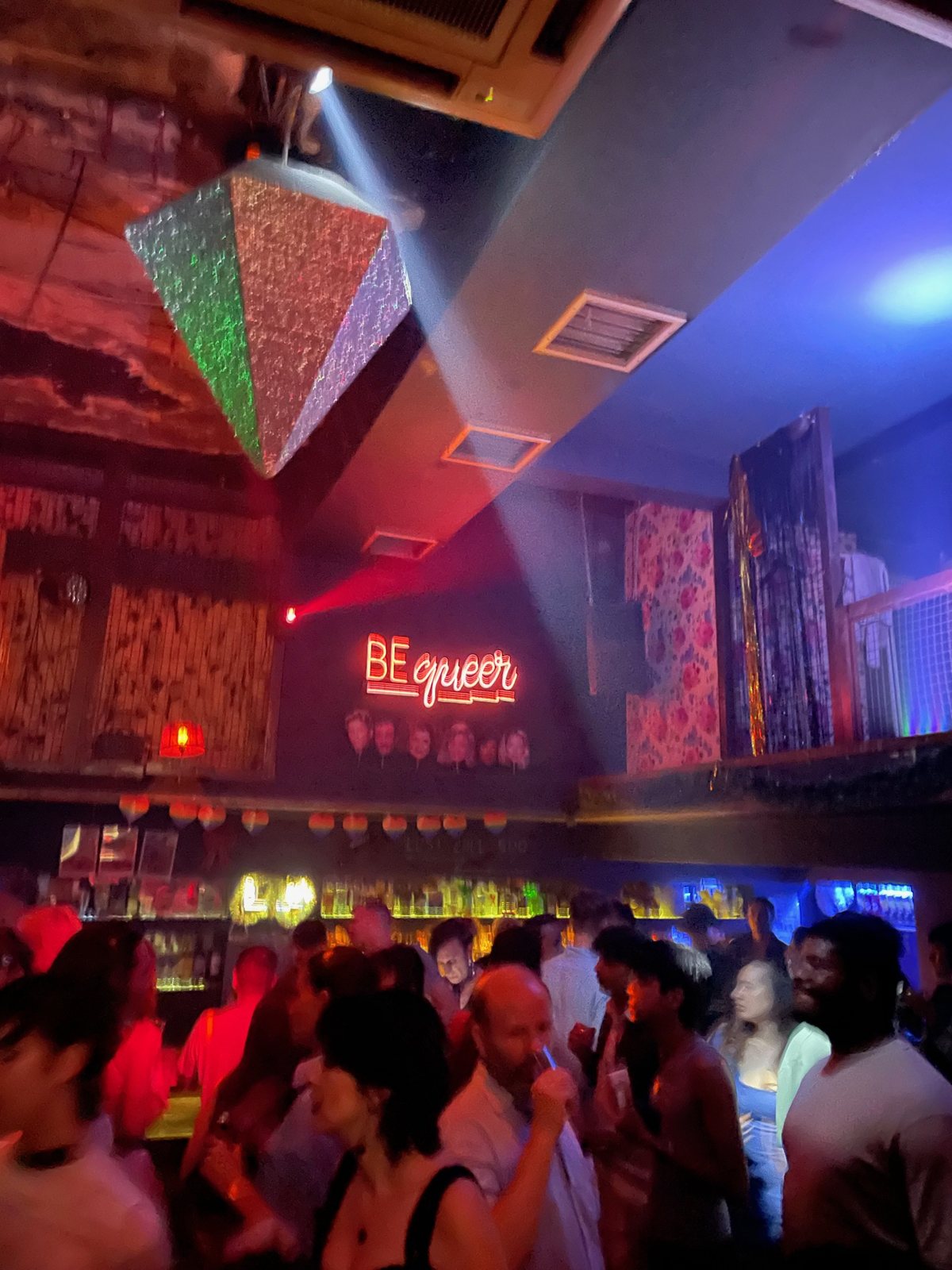 A crowded bar under coloured lights, a neon sign reading “Be queer” on the back wall.