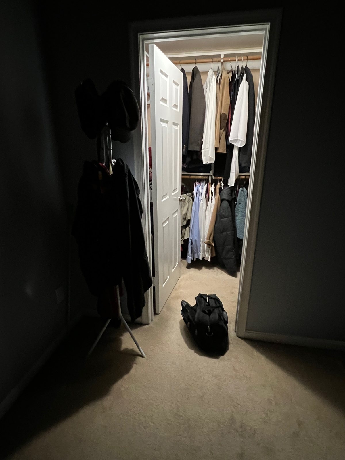 A lit wardrobe of hanging clothes seen from a dark hallway, a bag on the floor.