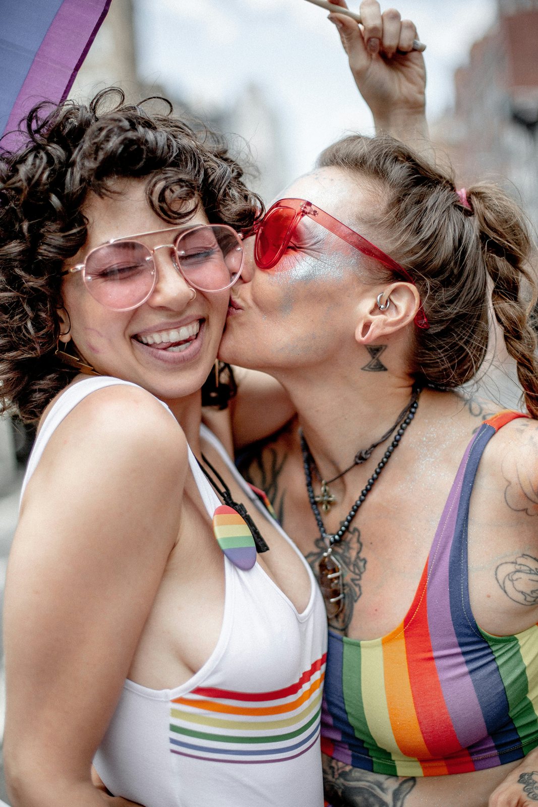 Two women in rainbow-striped tops and pink sunglasses, laughing, one kissing the other on the cheek.