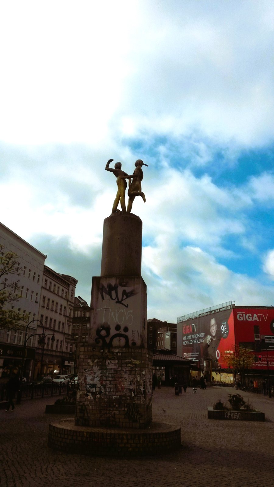 A graffitied stone monument topped by a statue of two figures, in a city square under bright cloud.